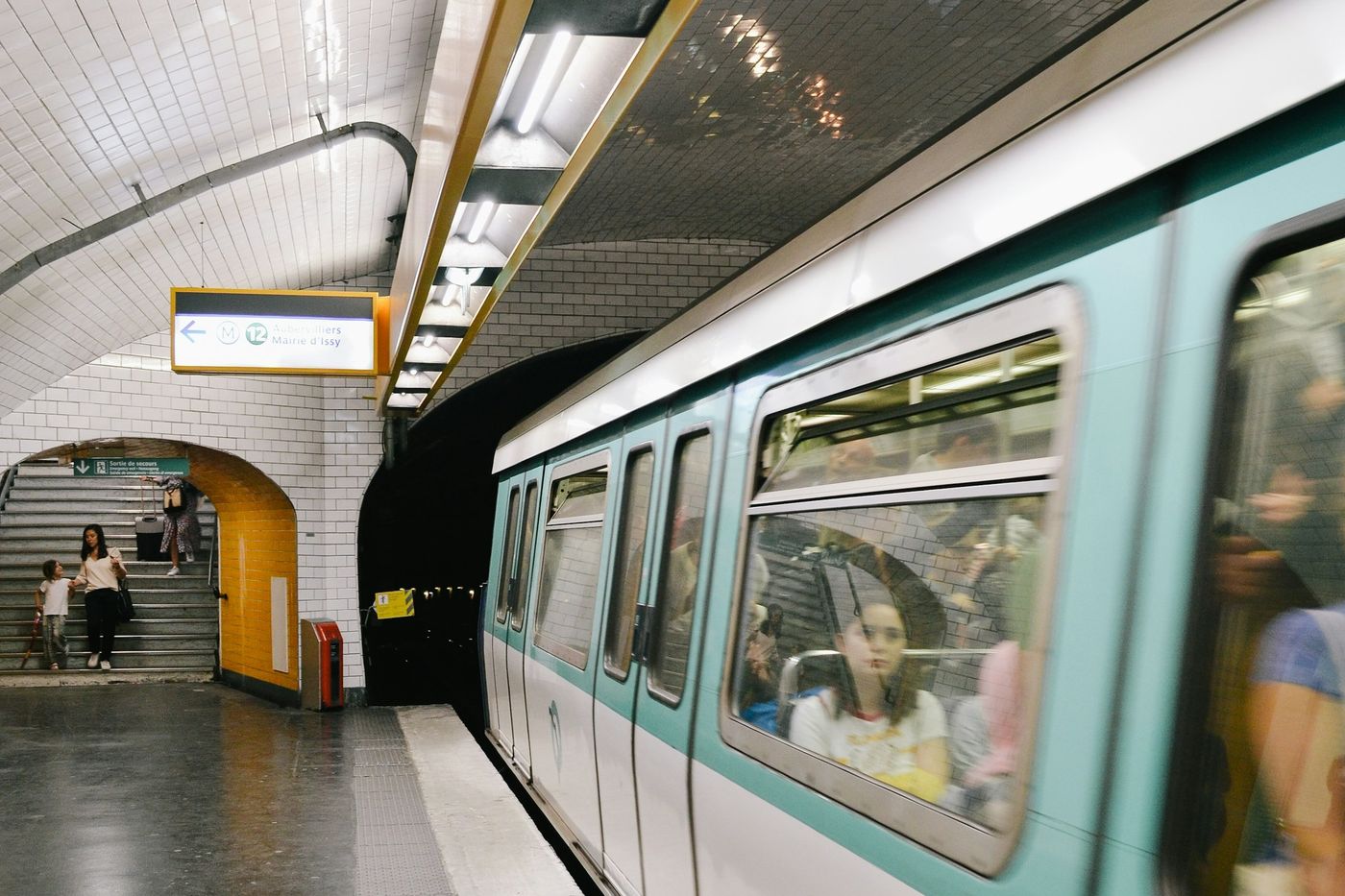 Quai de métro carrelé de blanc, une rame qui arrive et des voyageurs dans l'escalier du fond